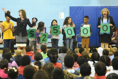 At an elementary school assembly in the gym, students and faculty hold up signs that spell out $25,000
