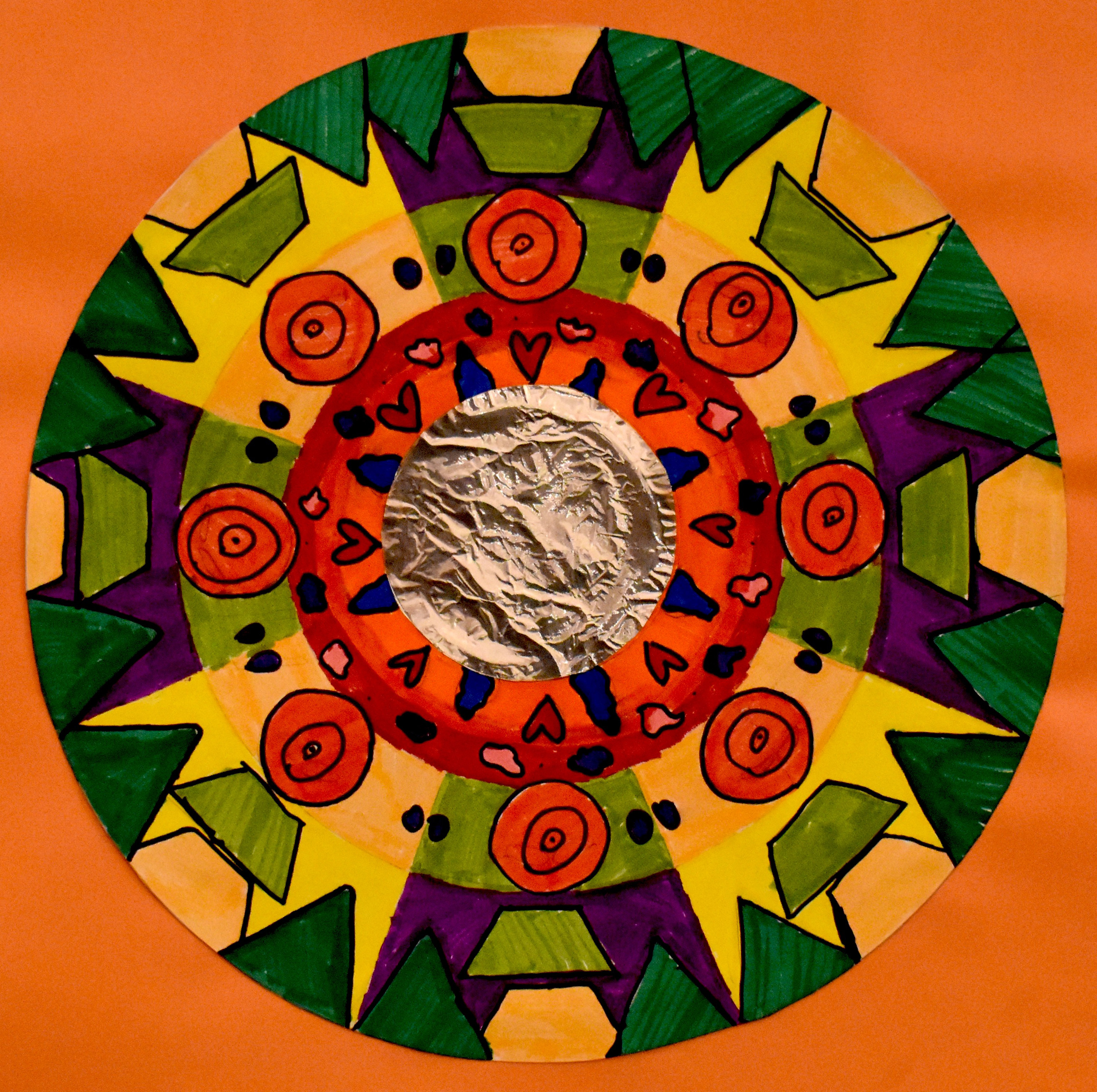 A colorful circular design with a metalic silver circle in the center, in green, purple, yellow, and red. Mixed media.